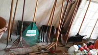 Photo showing multiple long-handled rakes, shovels, and garden tools leaning against a wall.