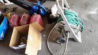 Wide view showing Kobalt electric blower, three red gas cans, garden hose on reel, and a box with lanterns.