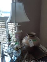Two clear glass oil lamps, a 32-inch tall white table lamp with shade, and a 14-inch ceramic vase on a glass table near window.