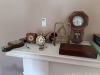 Vintage clocks of various sizes and styles including a wall-mounted Regulator A clock with pendulum, brass bell on stand, stereoscope, loupe, Weiss's Victorian medical kit box, and other small items displayed on a white shelf.