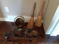Photo showing all items: brass and copper bucket, two fireplace brooms, wrought iron candle holder, and bellows placed on wood floor.