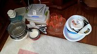 Wide shot showing all items grouped including plastic food containers, metal bowls and colander, plastic utensils, cups, plates and glass tray on floor