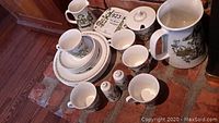 Full view of the dishware set arranged on a surface, showing pitchers, plates, cups, saucers, and lidded container with matching folk art design.
