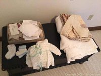 Photo showing folded white queen size cotton bedspread with infant crochet clothing laid out in front, including booties and crochet sweater, and a box of linens on top of black surface.