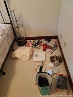 Photo showing layout of items including shower curtain, bathroom scale, curling irons, hair dryers, shaver, caddy, and office supplies on carpeted floor near bed.