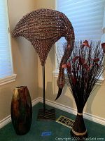 Three large decorative items grouped, showing the wood and straw ostrich sculpture with metal stand in the middle, multicolored glass vase on the left, and straw flowers on the right in corner setting.