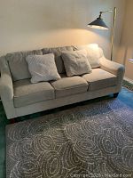 Full view of grey-beige sofa with three cushions, two throw pillows, floor lamp behind it and part of rug in front.