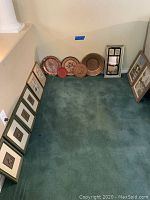 Overview of the lot showing framed artworks arranged around a corner and four decorative plates lined up on the floor.