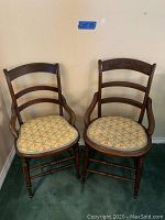 Two wooden side chairs with upholstered seats placed side by side against a beige wall, showing overall look and style of the chairs.