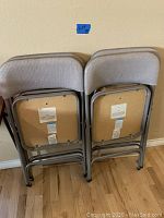 Four Samsonite folding chairs stacked against wall, showing padded backs and seats, beige upholstery, gray folding metal frames