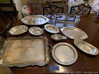Wide view of 10 silver plated serving platters and plates laid out on a table, showing varied shapes and sizes, some with engraved patterns