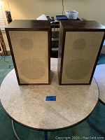 Front view of the two Smaller Advent Loudspeakers standing side by side on a round table, showing wooden cabinet and beige fabric covers.