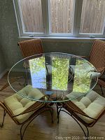 Glass round table with four wicker chairs arranged around it in front of a window with wooden blinds.