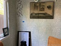 Photo showing a corner of the room with multiple framed artwork pieces: one large framed vintage-style print hanging on the wall and two smaller framed prints leaning against the wall near a window with natural light.