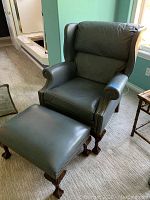 Blue gray faux leather wingback chair with ottoman seen from an angle next to a window and small side table