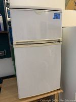 Front view of small white Frigidaire fridge with freezer compartment and handles, showing full closed unit.
