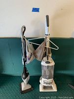 Two upright vacuums side by side against a green and white wall, with a hanger and plastic bags hanging off one vacuum.