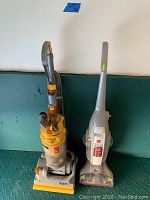 Full view of Dyson and Hoover upright vacuums side by side on floor against a wall, showing both models and some cords.