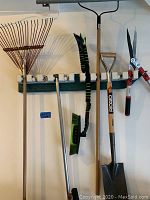 Photo showing a metal leaf rake, broom, shears, shovel, and part of a hose reel hung on wall hooks.