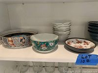 Shelf showing assorted bowls including a large decorative bowl with Asian floral pattern, a teal bowl with white flowers, white scalloped small bowls, and a white plate with orange floral pattern.