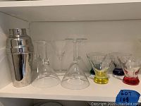 Shelf with stainless steel cocktail shaker, several clear glass martini glasses, some with colorful bases and stems in yellow, red, purple, and blue.