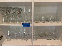 Shelf with 20 assorted clear glass wine glasses and tumblers, including ornately stemmed glasses.