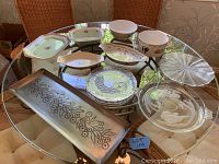 Wide shot of all ten serving dishes and bowls arranged on a glass table. Includes ceramic dishes with floral patterns, clear etched glass bowl with lid, clear glass segmented platter, and rectangular metal tray with embossed branch design.