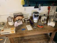 Wide photo showing full lot on wooden table including mixers, food processors, coffee maker, electric knife box, chafing dish box, utensils, bowls, and attachments