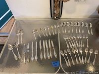 Full view of flatware laid out on glass table showing assortment of spoons, forks, knives and serving spoons, all new and unused.