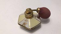Top view of vintage hexagonal perfume atomizer bottle showing cream and white panels with gold floral accents and attached brass spray bulb covered in mauve fabric.