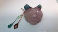 Blown glass cat mask with attached glass decorations shown laying flat on white surface. Mask is translucent pink with blue ears and three hanging glass pieces attached by multicolored strings.