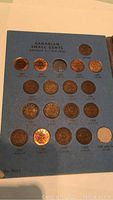 Close-up of blue booklet page titled 'Canadian Small Cents George V - 1910-1935' with 18 Canadian pennies inserted showing obverse and reverse sides with years ranging from 1920 to 1935.