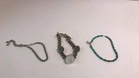 Three bracelets displayed on a white surface: silver chain bracelet on left, handmade quartz and iridescent stone bracelet center, turquoise beaded bracelet right.