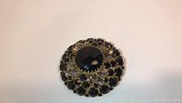 Top-down image of round black rhinestone brooch or pendant with a large central black crystal and multiple smaller black rhinestones in two concentric rings.