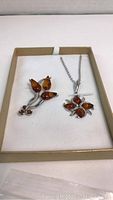 Flower-shaped amber pendant on rhodium plated chain along with matching amber brooch inside jewelry box on white cushion.