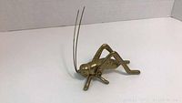 Brass grasshopper figurine viewed at an angle showing the head, detailed legs, and long thin antennae.