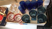 Photo showing a variety of clear crystal glasses, cobalt blue tall glasses, blue patterned wine glasses, and small clear glasses with red rims arranged in a box.