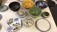 Photo showing group of various pottery bowls and plates with floral, solid color, and scalloped designs arranged on a table.