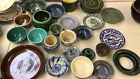 Top-down view showing whole pottery assortment on white surface including green, blue, brown, and cream bowls plus a large dark brown bowl and blue and white decorative plate.