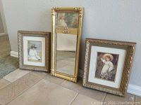 Three framed pieces: two artwork prints and one tall framed mirror with artwork on top, arranged on floor against wall.