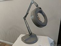 Full view of gray illuminated magnifying glass lamp showing flexible arm and weighted base on table.