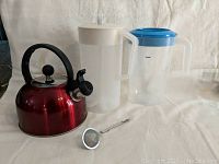 Red whistling tea kettle, two plastic pitchers, and metal tea strainer placed together for reference