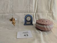 Set of three items on fabric backdrop: blue Wedgwood clock, rose-colored potpourri holder, clear glass bell with angel inside