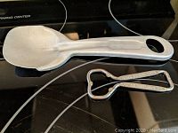 Kentucky Fried Chicken ice cream scoop and Potosi Beer Company bottle opener side by side on a black stovetop surface, showing the scoop's metal construction and flat design with a hole in the handle, and the bottle opener's classic shape.