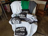 Pile of assorted Colorado State University men's shirts in various sizes and colors including white, gray, green, and black arranged on a chair