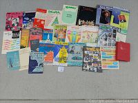 Overview photo showing the full assortment of about 30 vintage sheet music booklets spread out on a floor, showcasing a colorful variety of covers and titles from musicals and genres.