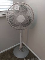 White Holmes standing fan with circular grille and round base, shown plugged in and positioned near a window and wall corner.
