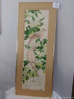 Framed batik painting showing two doves perched among green leaves and white flowers, with matting and a golden frame, vertical orientation.