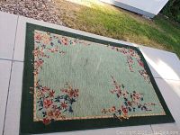 Full view of the large rectangular area rug outdoors on a paved surface and grass, showing the floral design and green border.