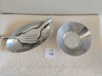 Two Nambe Ware salad bowls and serving utensils on white cloth: one oval bowl, one smaller bowl with patina, one serving fork and one serving spoon.